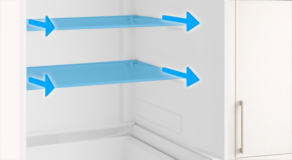 Wnętrze lodówki z dwoma półkami oznaczonymi niebieskimi strzałkami wskazującymi możliwość regulacji. Obok widoczna jest część białej szafki z metalowym uchwytem.