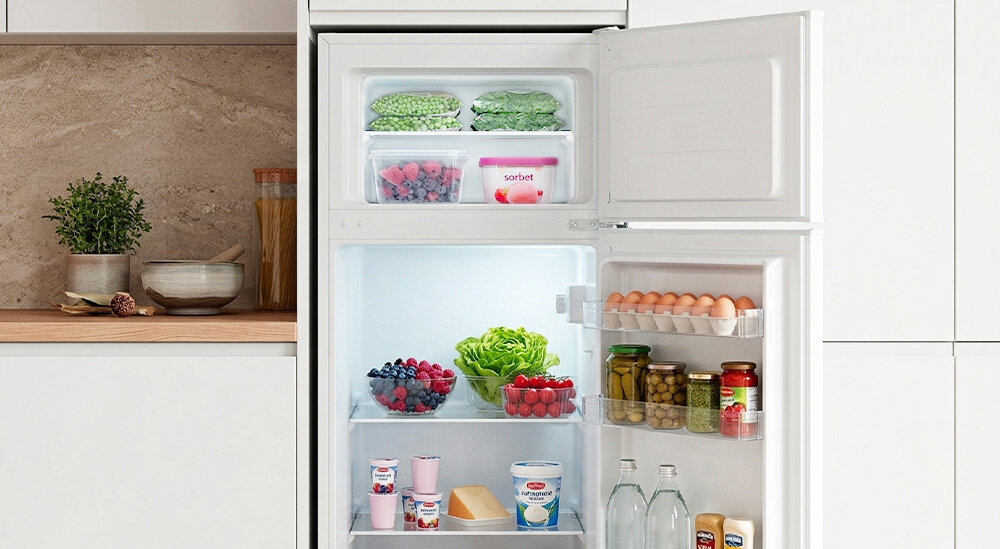 Otwarta lodówka z widocznymi produktami spożywczymi, takimi jak warzywa, owoce, jajka, napoje i jogurty. Na górnej półce zamrażarki znajdują się mrożonki i pojemnik z napisem \'sorbet\'.