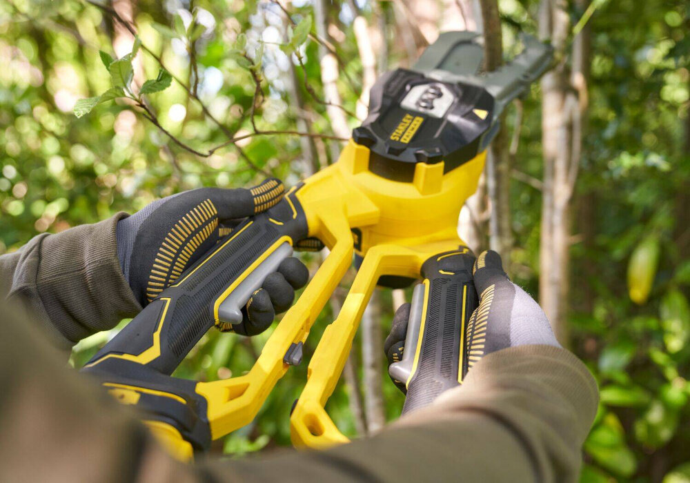 Sekator do gałęzi akumulatorowy STANLEY SFMCPS515B-XJ ogród zbliżenie na uchwyty osoba trzyma sekator w dłoniach w tle gałęzie podwójne uchwyty z przełącznikami wymagają użycia obu rąk do uruchomienia sekatora bezpieczeństwo użytkownika kontrolować każdy ruch ogranicza ryzyko przypadkowego włączenia ułatwia stabilne prowadzenie cięcia