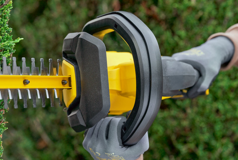 Nożyce do żywopłotu STANLEY SFMCHT655B-XJ 550mm 20V akumulatorowe ogród osoba trzyma nożyce w dłoniach zbliżenie na uchwyt wymagają użycia obu rąk do uruchomienia nożyc minimalizuje ryzyko przypadkowego włączenia ostrza bezpieczeństwo użytkownika