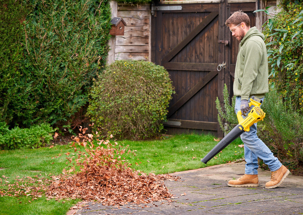 Dmuchawa do liści akumulatorowa STANLEY 20V SFMCBL714B-XJ SOLO ogród osoba zdmuchuje liście z chodnika w tle krzewy brama wjazdowa porządek zamiatanie czyszczenie wokół domu