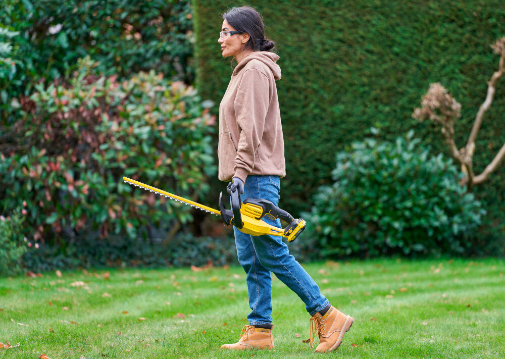 Nożyce do żywopłotu STANLEY SFMCHT650D1-QW 500mm 20V akumulatorowe ogród osoba idzie po trawniku z nożycami w dłoni w tle rośliny waga około 2,9 kg zmniejsza obciążenie ramion użytkownik odczuwa większy komfort