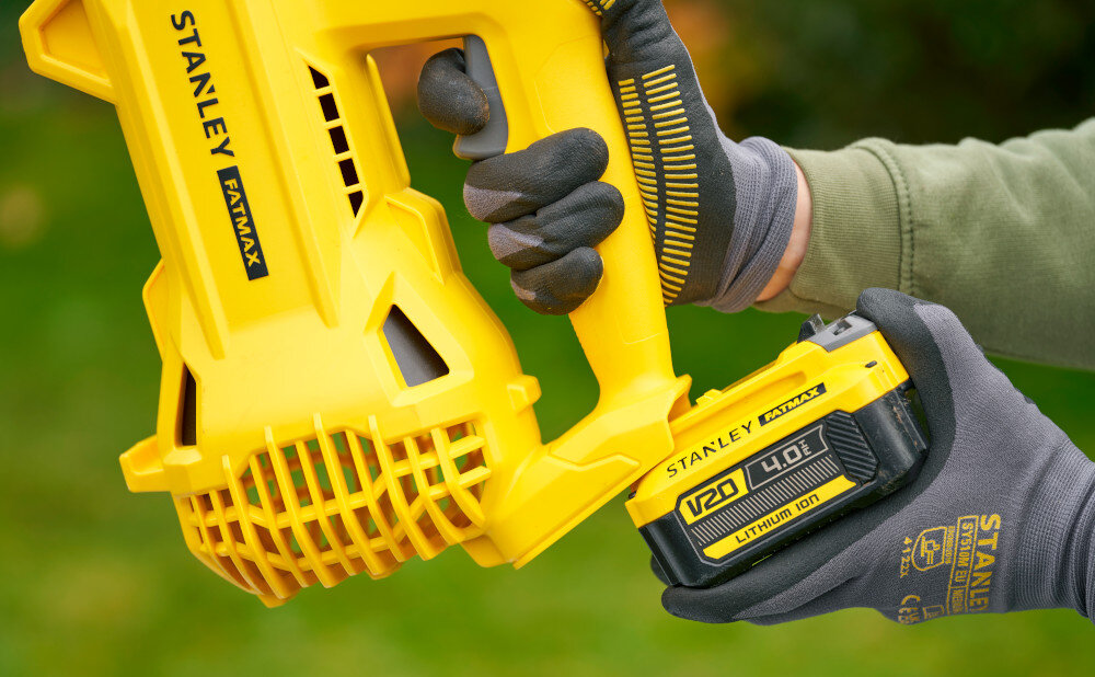 Osoba trzyma żółtą dmuchawę do liści marki STANLEY FATMAX z akumulatorem V20 4.0Ah Lithium Ion. Widoczne są rękawice ochronne na dłoniach osoby obsługującej urządzenie.