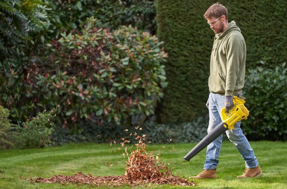 Osoba używa żółtej dmuchawy do liści na trawniku, kierując strumień powietrza na stos suchych liści. W tle widoczne są krzewy i żywopłot.