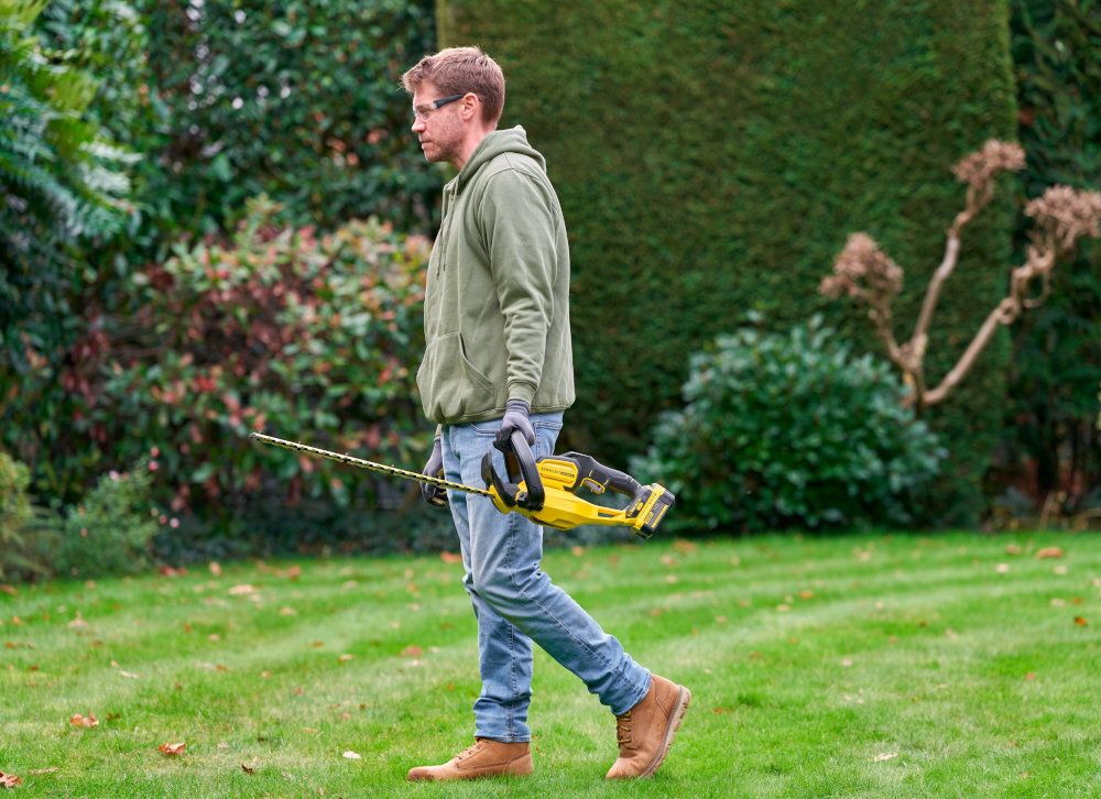 Nożyce do żywopłotu STANLEY SFMCHT655M1-QW 550mm 20V akumulatorowe ogród osoba idzie po trawniku z nożycami w dłoni w tle rośliny waga około 2,9 kg zmniejsza obciążenie ramion użytkownik odczuwa większy komfort