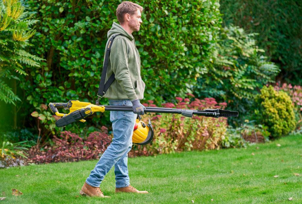 Piła akumulatorowa STANLEY SFMCPS720B-XJ SOLO ogród osoba idzie po trawniku w dłoni trzyma piłę w tle drzewa krzewy rośliny silnik bezszczotkowy płynna i przewidywalna praca kontrola nad narzędziem precyzyjne prowadzenie cięcia i ogranicza szarpanie gałęzi