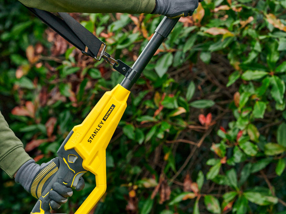 Piła akumulatorowa STANLEY SFMCPS720B-XJ SOLO ogród osoba trzyma piłę w dłoniach zbliżenie na uchwyt i pasek w tle liście żywopłotu miękki uchwyt na kiju regulowany pasek na ramię komfort pracy waga około 3,6 kg stabilne podparcie ułatwia precyzyjne prowadzenie narzędzia