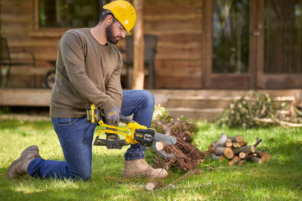 Sekator do gałęzi akumulatorowy STANLEY SFMCPS515M1-QW ogród trawnik osoba tnie gałęzie klęcząc na trawniku w tle drewniany dom przycinanie drzew cięcie powalonych gałęzi na mniejsze kawałki szybkie i precyzyjne formowanie roślin ergonomicznemu kształtowi