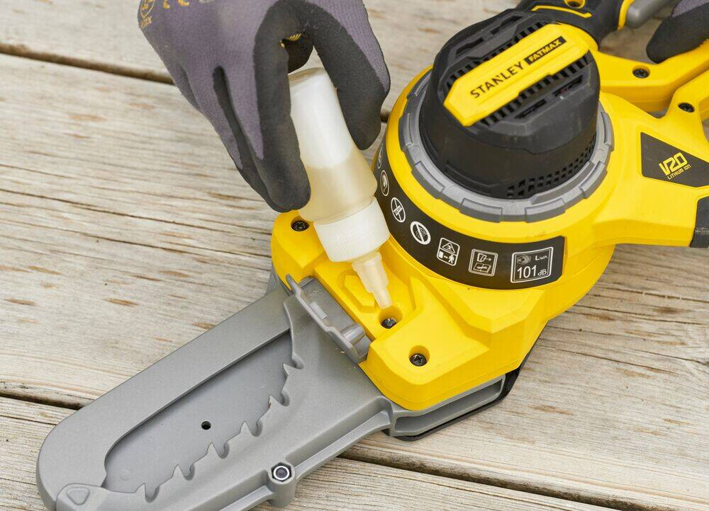 Sekator do gałęzi akumulatorowy STANLEY SFMCPS515M1-QW drewniany blat osoba przykłada buteleczkę z olejem do sekatora system półautomatycznego smarowania regularnie dostarcza olej do łańcucha zmniejsza tarcie i zużycie narzędzie zachowuje wysoką wydajność cięcie jest płynne ryzyko zacięć i uszkodzeń elementów tnących jest niskie Półautomatyczne smarowanie przekłada się na długą żywotność