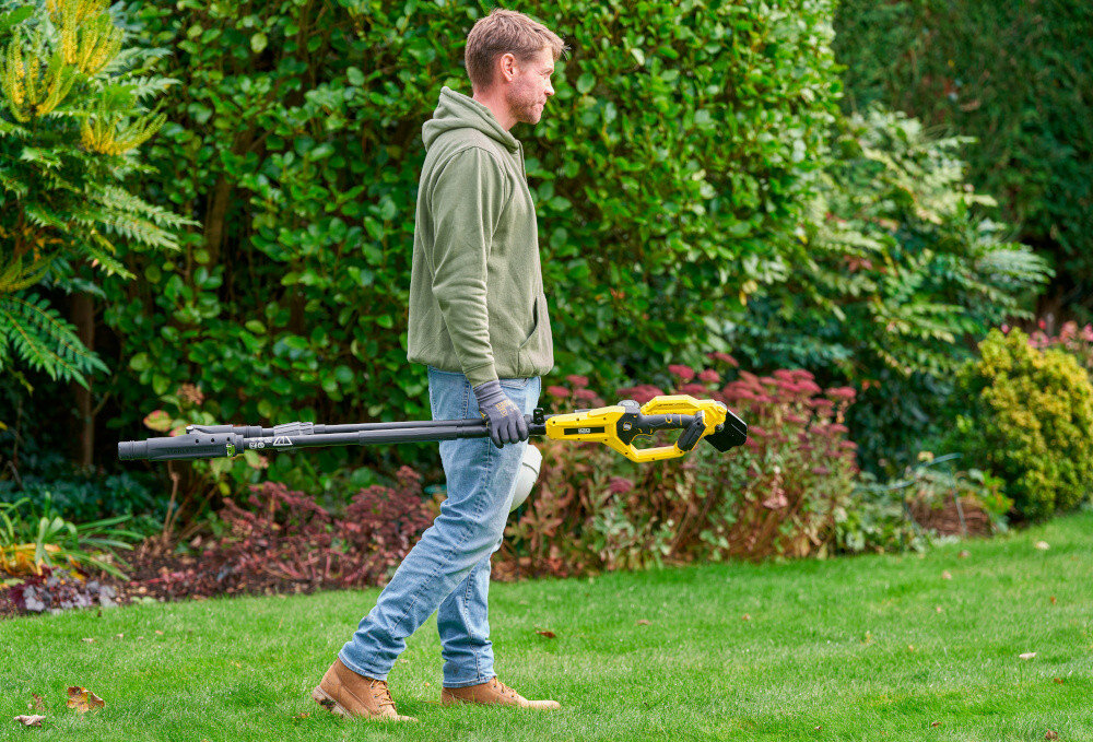 Sekator do gałęzi akumulatorowy STANLEY FatMax SFMCPP632D1-QW osoba idzie po trawniku w dłoni trzyma złożony sekator w tle krzewy drzewa  miękki uchwyt na tyczce poprawia wygodę użytkowania pewniejszy chwyt wadze około 3,62 kg stabilne i łatwe w prowadzeniu ogranicza zmęczenie ramion