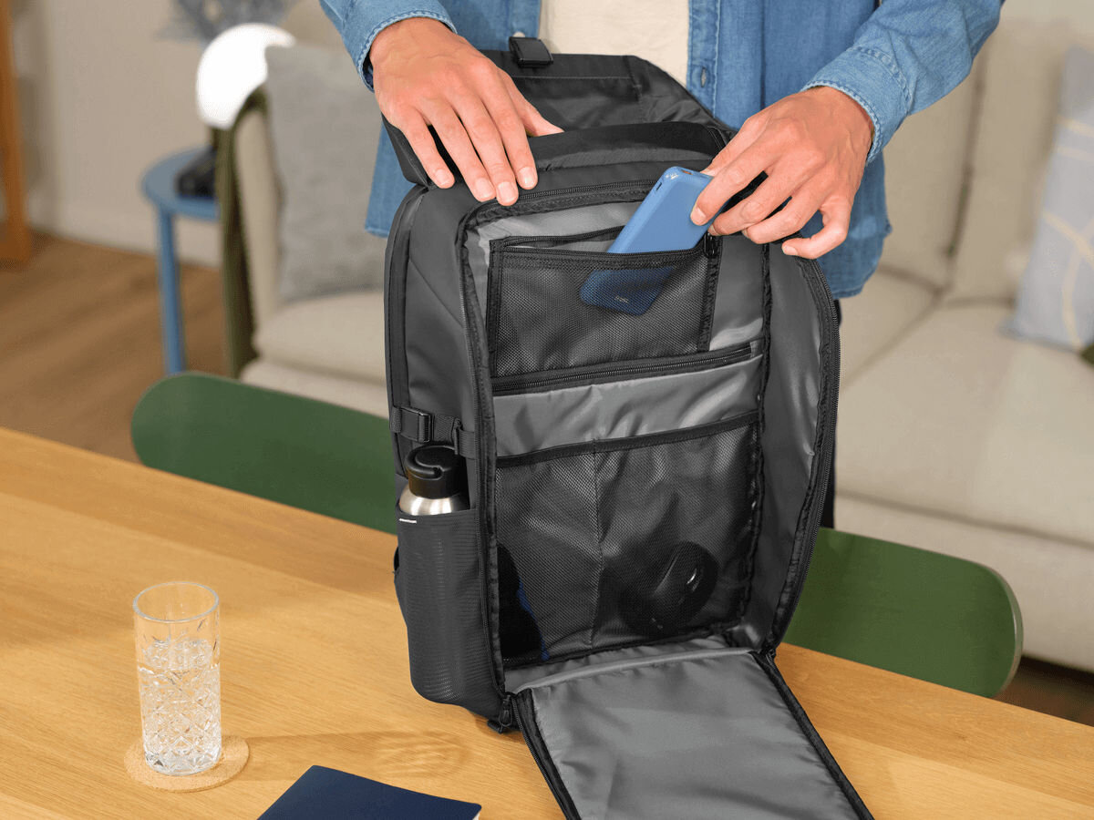 Plecak na laptopa TRUST Jersey 17 cali Czarny Zbliżenie na otwarty czarny plecak Trust, ukazujące wnętrze. Plecak ma liczne kieszenie organizacyjne, w tym siatkowe. Ręce mężczyzny wkładają niebieski smartfon do jednej z małych kieszeni.