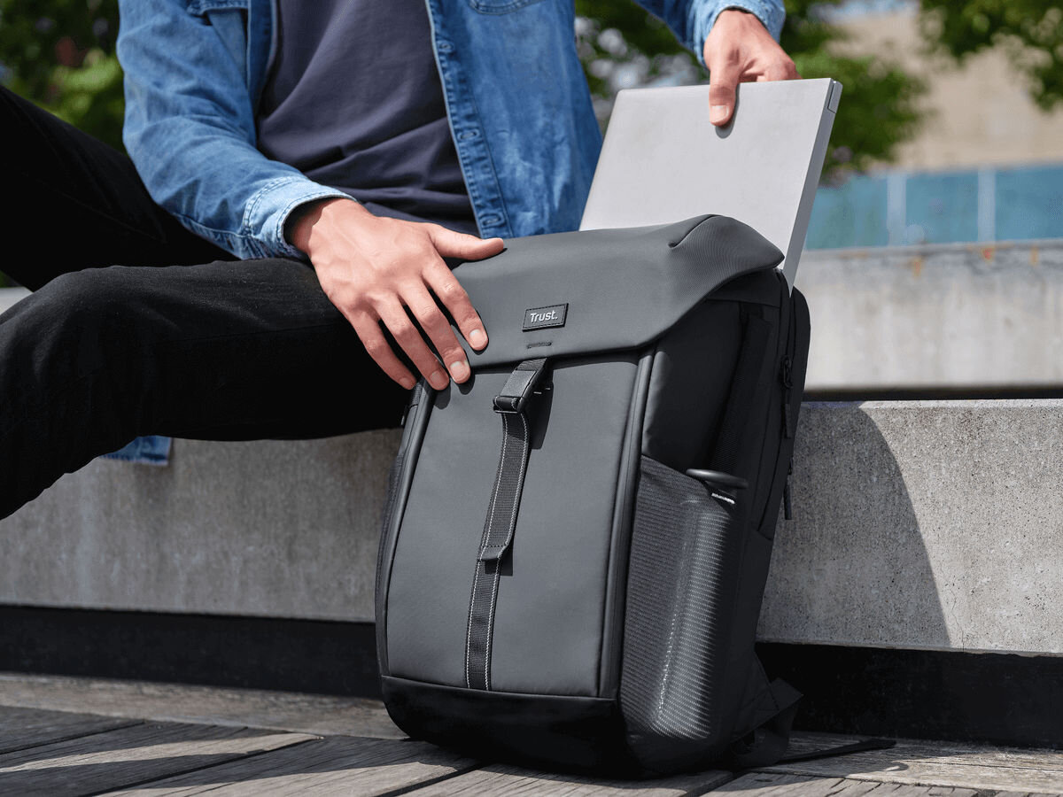 Plecak na laptopa TRUST Jersey 17 cali Czarny Mężczyzna, siedzący na betonowej ławce, otwiera górną klapę czarnego plecaka Trust i wkłada lub wyjmuje z głównej komory szary laptop.