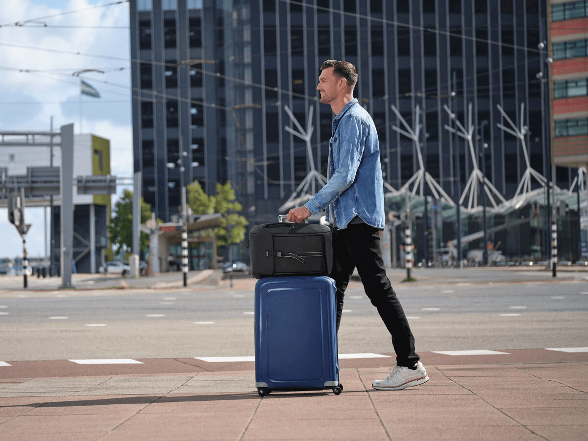 Plecak na laptopa TRUST Jersey 17 cali Czarny Mężczyzna w dżinsowej koszuli i czarnych spodniach uśmiecha się, ciągnąc niebieską walizkę na kółkach, na której leży czarna torba podróżna. Idzie po chodniku obok nowoczesnego, dużego budynku biurowego.