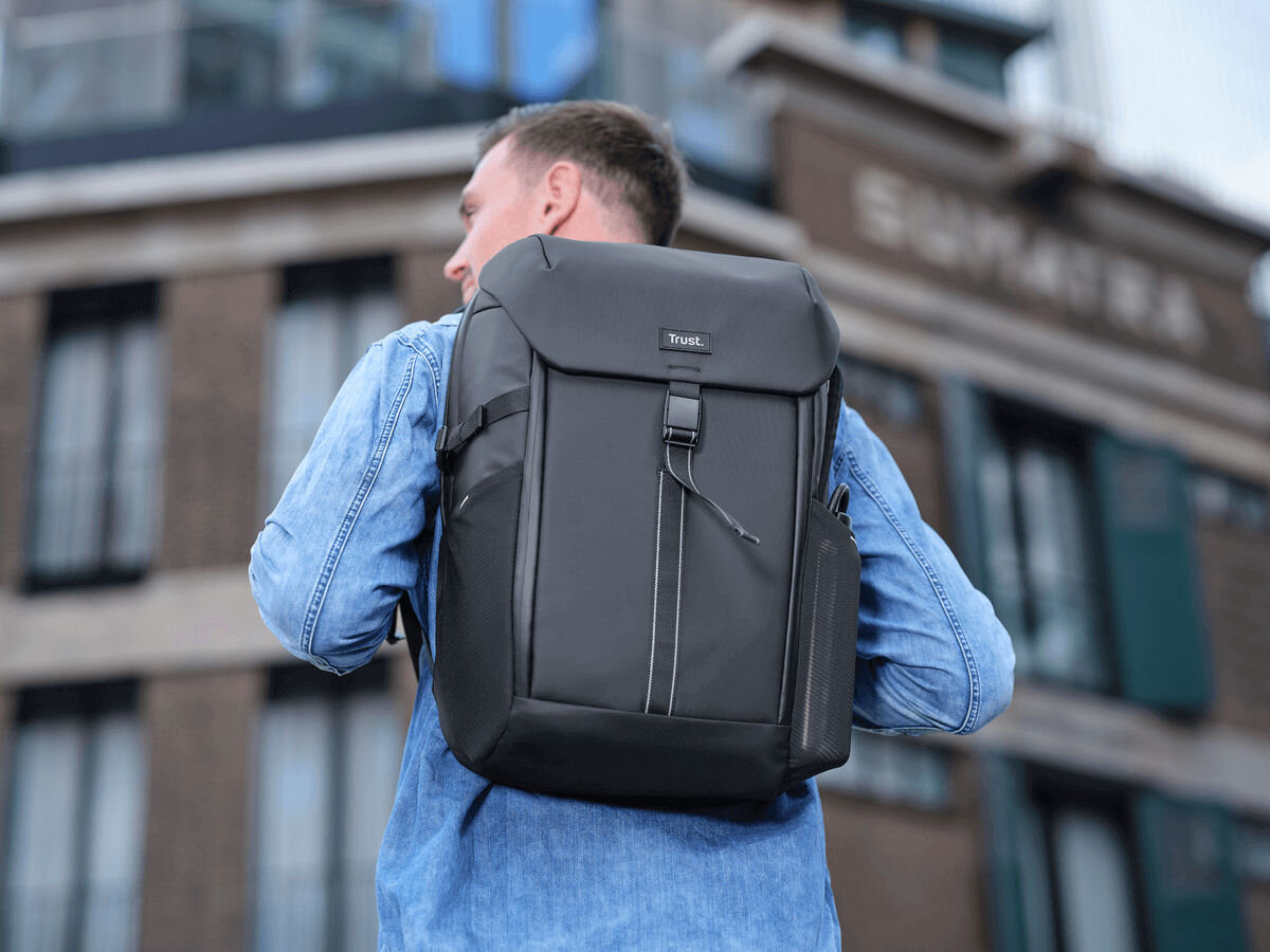 Plecak na laptopa TRUST Jersey 17 cali Czarny Mężczyzna ubrany w dżinsową koszulę i czarny plecak Trust (widok z tyłu/boku) stoi na ulicy przed ceglanym budynkiem, gotowy do drogi.