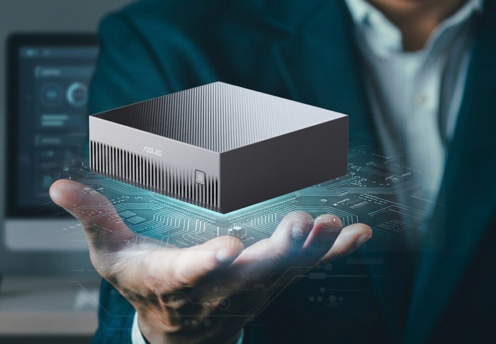 Komputer ASUS Ascent GX10 Srebrny komputer typu mini PC lewitujący nad otwartą dłonią osoby w ciemnej marynarce, z nałożoną grafiką świecących, błękitnych ścieżek układu scalonego pod urządzeniem oraz rozmytym monitorem w tle