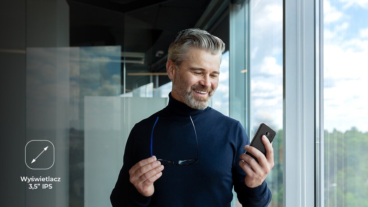 Mężczyzna stojący przy oknie w uśmiecha się, trzymając w dłoni telefon myPhone 3510 LTE i okulary; ikona wyświetlacza 3,5.