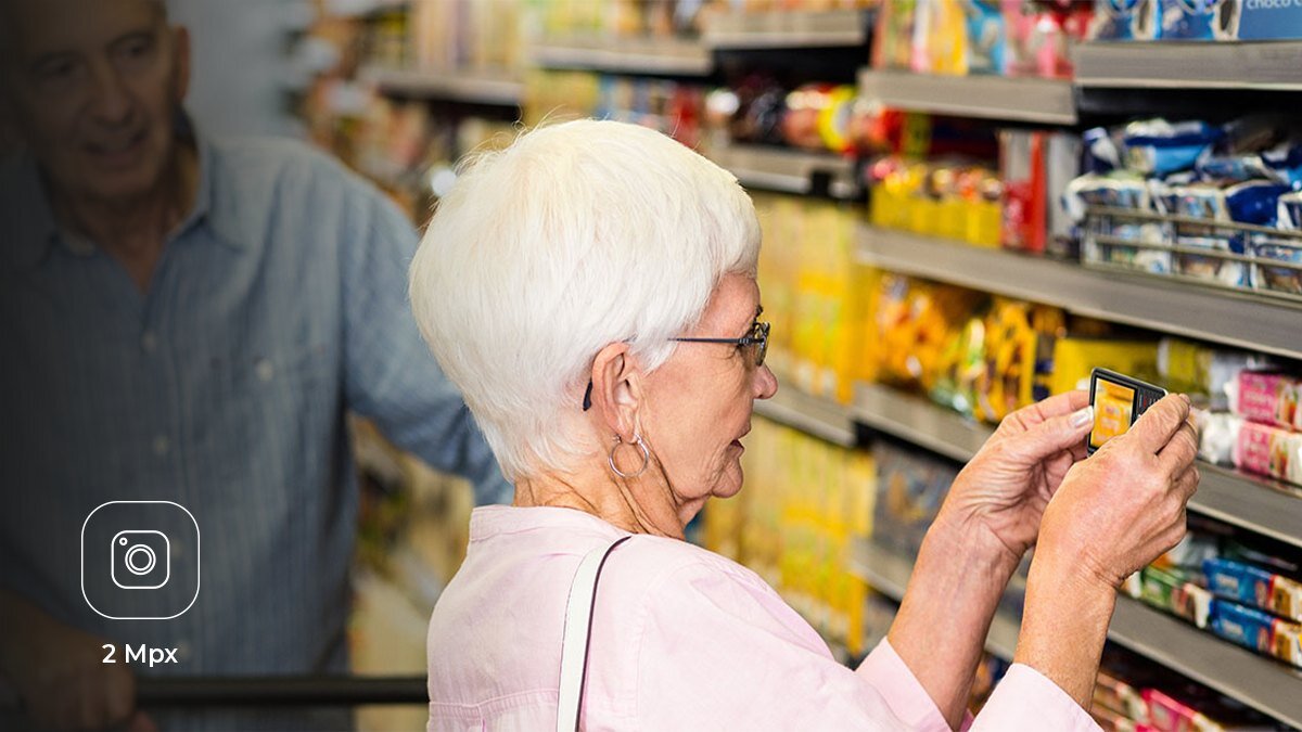 Zdjęcie lifestyle do myPhone 3510 LTE - starsza kobieta w markecie fotografuje etykietę na półce telefonem, w tle starszy mężczyzna z wózkiem; ikona aparatu.
