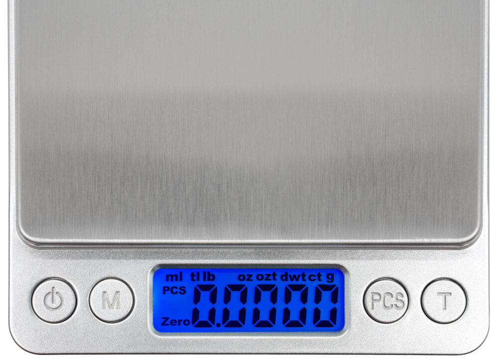 Waga jubilerska z metalową powierzchnią ważącą i niebieskim podświetlanym wyświetlaczem LCD pokazującym \'0.0000\'. Na panelu znajdują się przyciski oznaczone symbolami: włącznik, \'M\', \'PCS\', \'T\'.