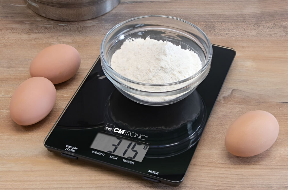 Waga kuchenna CLATRONIC KW 3626 Czarny precyzyjne ważenie czarna waga na blacie mąka w misce na wadze obok leżą jajka wyświetlacz