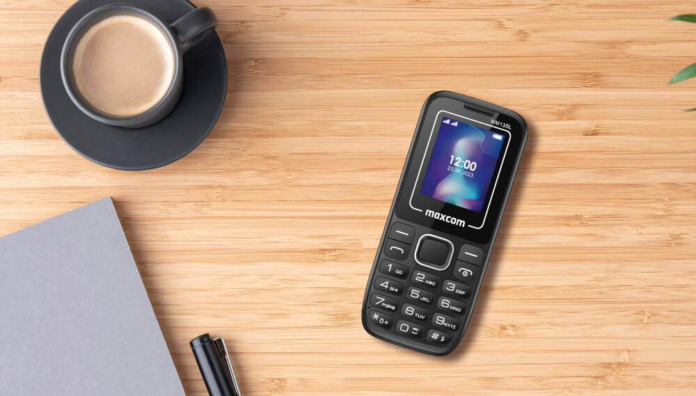 Na drewnianym blacie leży czarno-niebieski telefon z klawiaturą, na ekranie widoczna jest godzina 12:00. Obok telefonu znajduje się filiżanka kawy, notes i długopis.