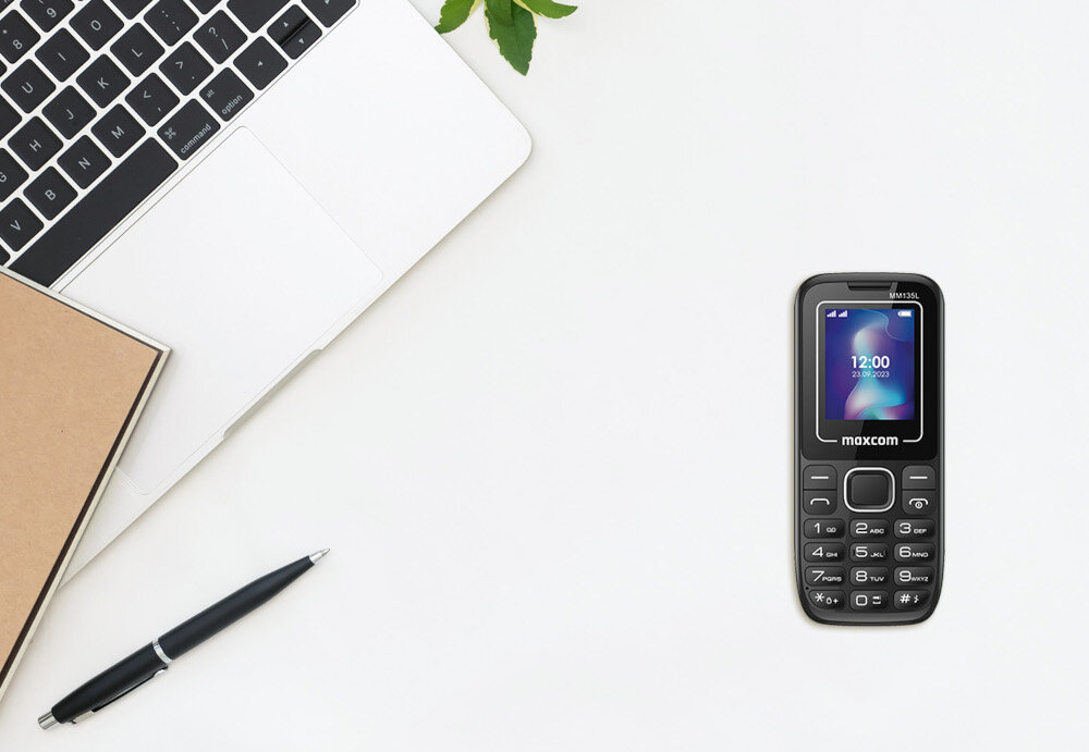 Na białym biurku znajduje się czarno-niebieski telefon komórkowy z wyświetlaczem pokazującym godzinę 12:00 i datę 23.09.2023. Obok telefonu leży otwarty laptop, notes i długopis.