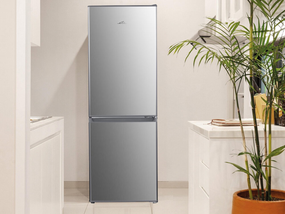 Lodówka ETA ETA385690010C Lodówka zamknięta w stalowym kolorze wkomponowana w jasną, nowoczesną kuchnię z rośliną w donicy na pierwszym planie