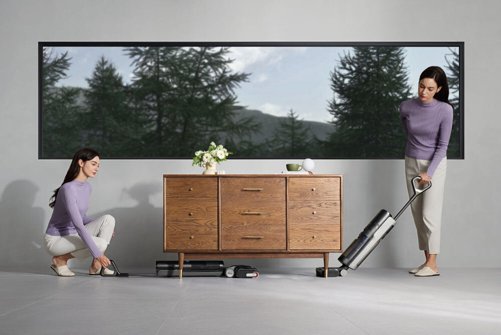 Dwie osoby używają odkurzacza w nowoczesnym wnętrzu z drewnianą komodą i dużym oknem z widokiem na drzewa. Na komodzie znajdują się kwiaty i filiżanka.