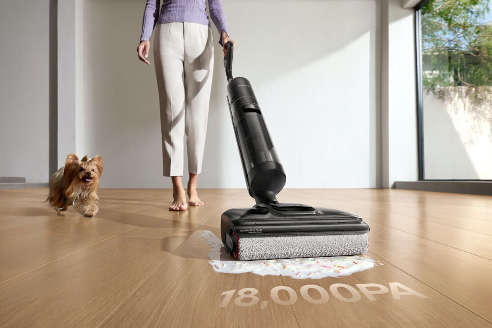 Osoba używa czarnego odkurzacza na drewnianej podłodze, obok znajduje się pies. Na podłodze widoczny jest napis \'18,000PA\'.