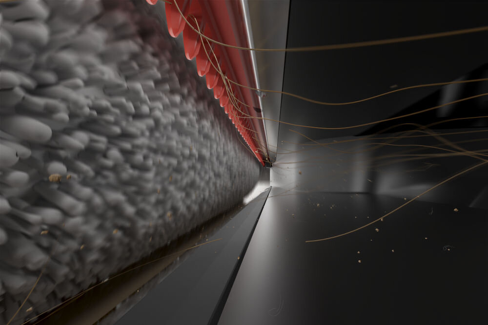 Zbliżenie na szczotkę odkurzacza z czerwonymi i szarymi elementami, zbierającą kurz i włosy z ciemnej powierzchni. Widać unoszące się drobinki kurzu i włosy.