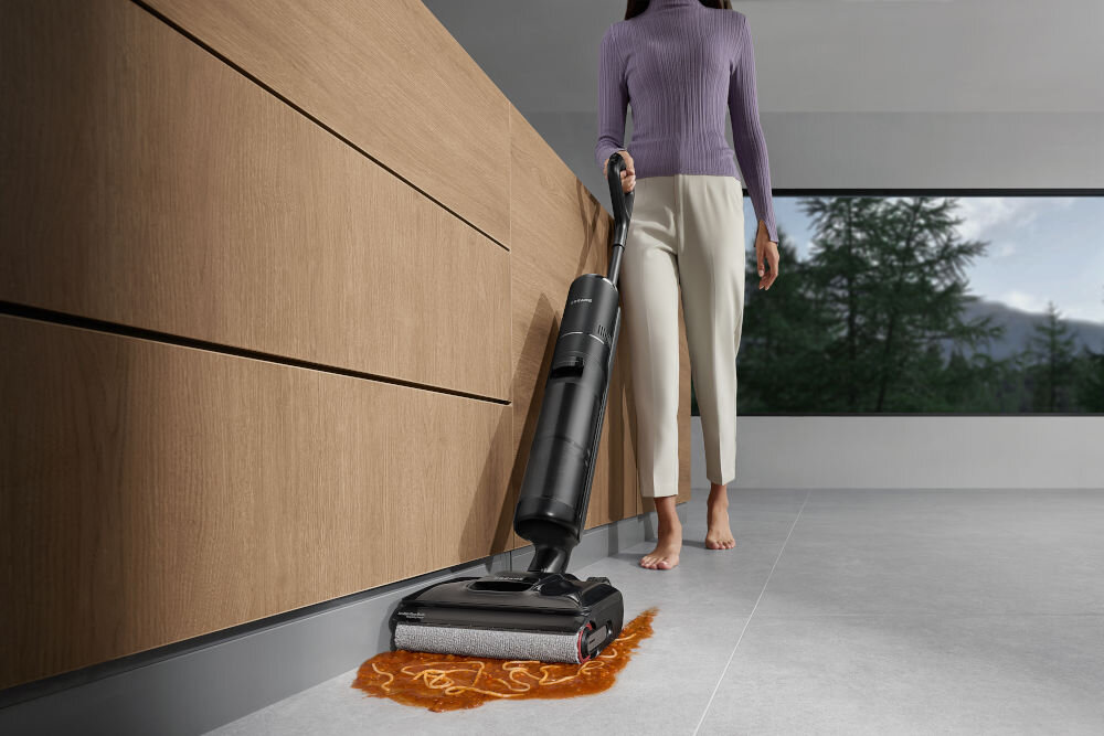 Osoba używa czarnego odkurzacza pionowego do czyszczenia rozlanego płynu na podłodze w nowoczesnym wnętrzu. W tle widoczne są duże okna z widokiem na drzewa i góry.