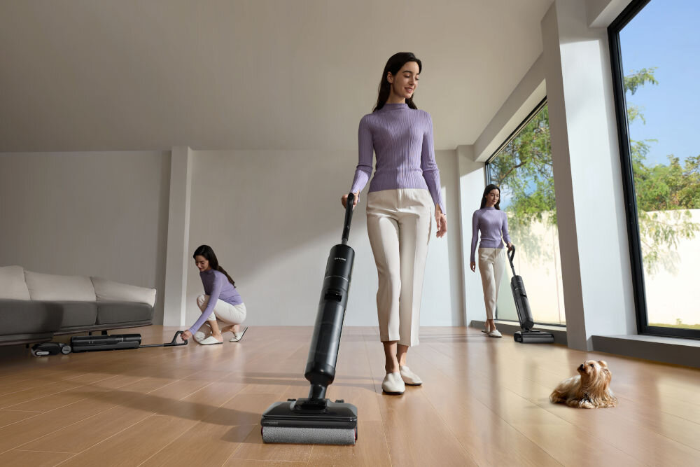 Trzy kobiety w fioletowych swetrach i białych spodniach używają odkurzacza na drewnianej podłodze w jasnym pomieszczeniu. Na podłodze siedzi mały piesek.