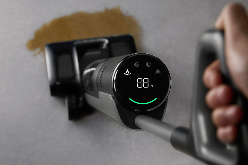 Osoba trzyma odkurzacz z wyświetlaczem pokazującym 88% i ikonami. W tle widoczna jest rozlana substancja na podłodze.