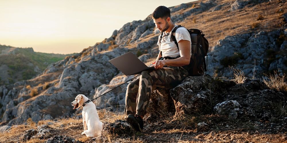 Mężczyzna z plecakiem pracujący na laptopie ASUS ProArt P16 siedząc na skalistym wzgórzu, obok biały pies, krajobraz górski w tle podczas złotej godziny - <span>ProArt P16</span> zaprojektowano z myślą o wygodnej mobilności i trwałości 