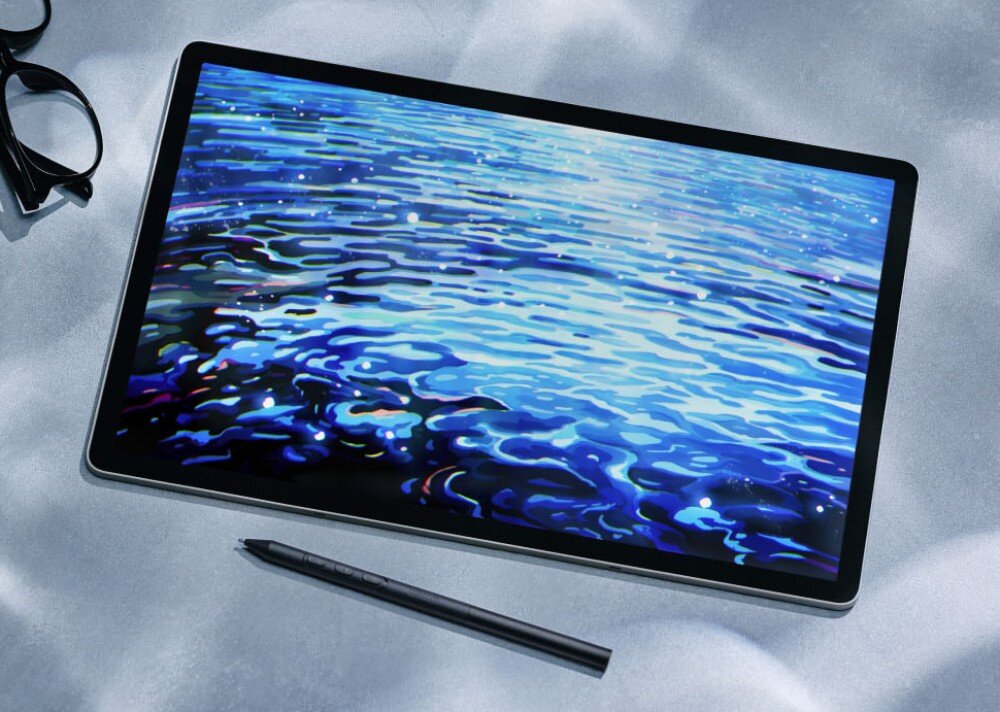 Na obrazie widać tablet graficzny z wyświetlaczem przedstawiającym abstrakcyjne fale w odcieniach niebieskiego. Obok tabletu leży rysik oraz okulary.