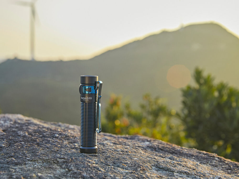 LATARKA OLIGHT BATON 3 PRO CW 1500LM 175M 120 DNI, Ergonomiczna, moc, lumeny, zasięg, dioda, latarka stoi pionowo na skale w tle teren górzysty