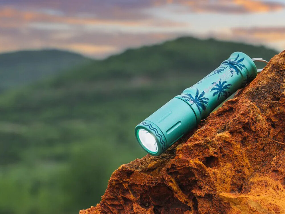 Latarka OLIGHT I3E EOS OL692821 Letnie Palmy Zielony, miniaturowa, jasność, 90 lumenów, codzienne, Edycja specjalna Olight I3E EOS Palmy leży na skale, tło rozmyte zielone wzgórza, zachód słońca.
