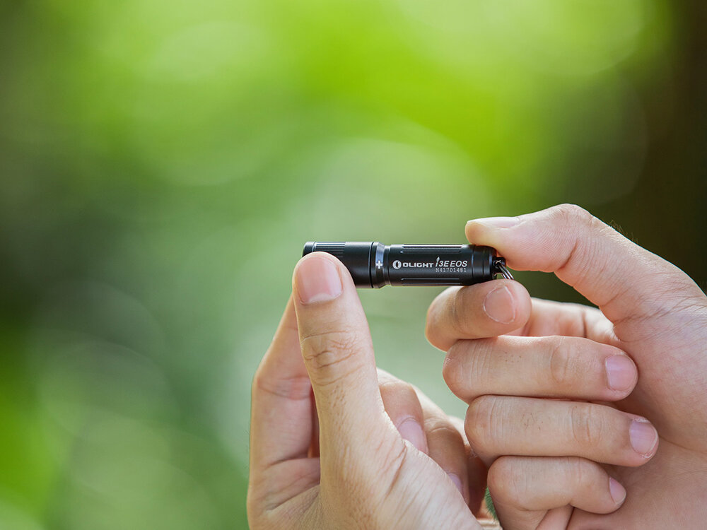 Latarka OLIGHT I3E-BK OL120137 Czarny, zasilanie, bateria, dostępność, czas, Czarna latarka Olight I3E EOS trzymana w dwóch dłoniach, zielone rozmyte tło w plenerze.