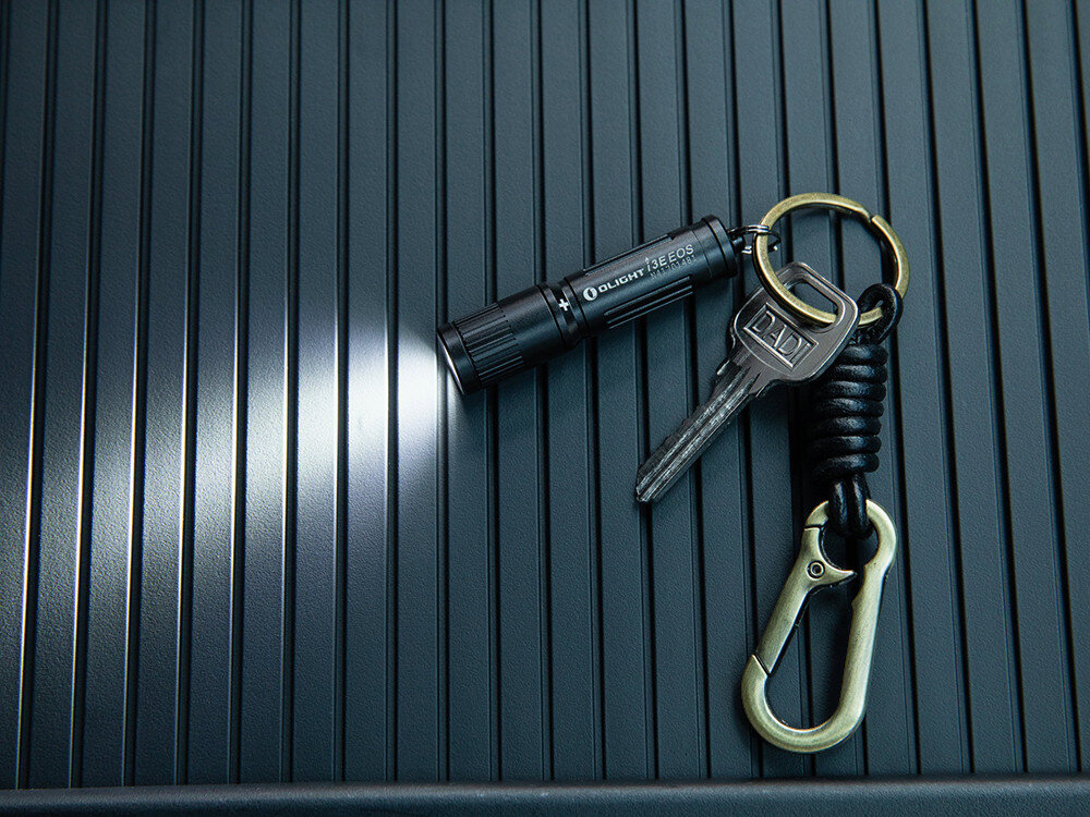 Latarka OLIGHT I3E EOS OL691510 Niebieski, szybkie, przekręcenie, głowica, bezpieczne, Latarka Olight I3E EOS z brelokiem i kluczem świeci na czarnym, żebrowanym tle, detal.