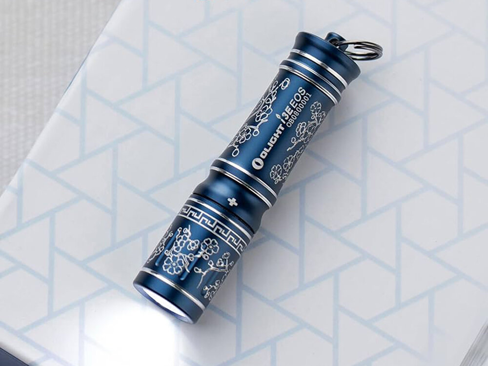 Latarka OLIGHT I3E EOS OL691510 Niebieski, zasilanie, bateria, dostępność, czas, Olight I3E EOS Sakura Blue zapalona na pudełku, eleganckie grawerowane kwiaty, białe geometryczne tło.