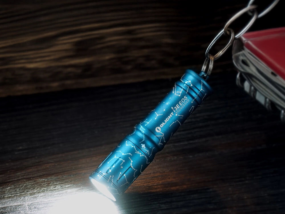 Latarka OLIGHT I3E EOS OL693712 Astral Cobalt Niebieski, zasilanie, bateria, dostępność, czas, Latarka Olight I3E EOS Constellation na kółku z kluczami, świeci białym światłem, ciemny blat.