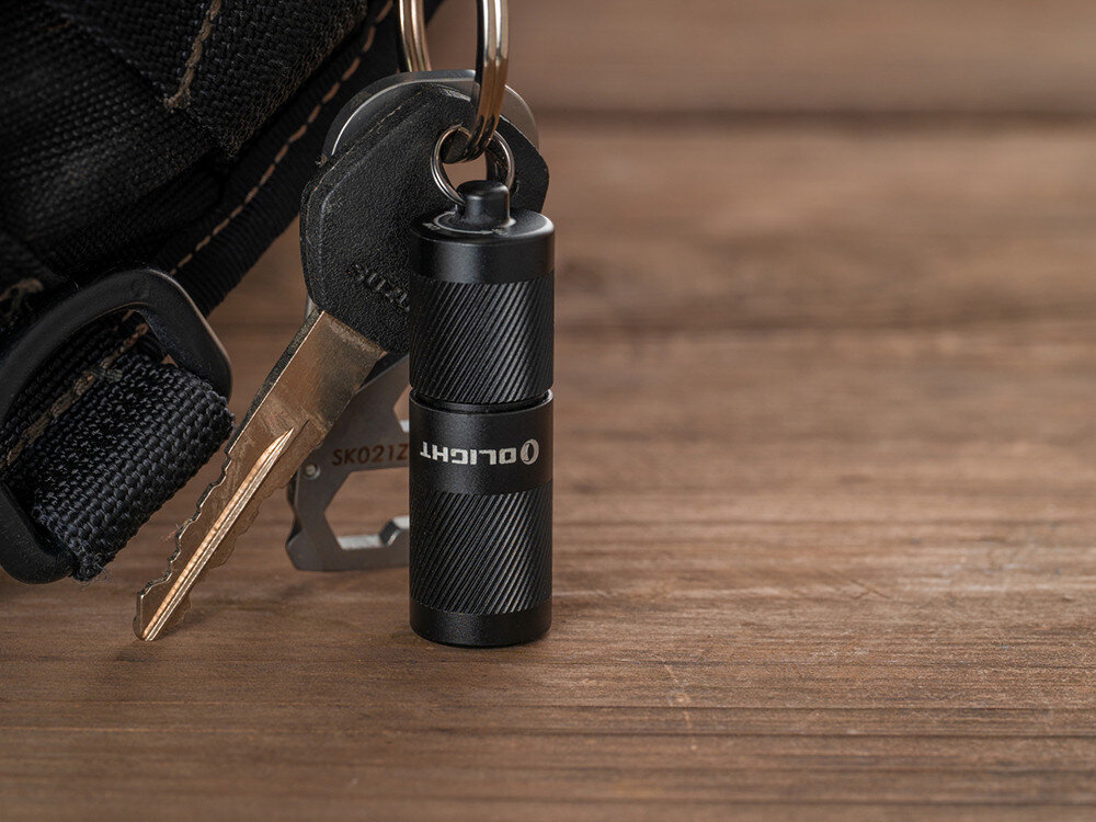Latarka OLIGHT I1R II EOS Kit, dwa tryby, 5 lumenów, 150 lumenów, obrót, Latarka Olight I1R 2 EOS przy kluczach, plecaku, klucz na drewnianym tle, minimalistyczny design.