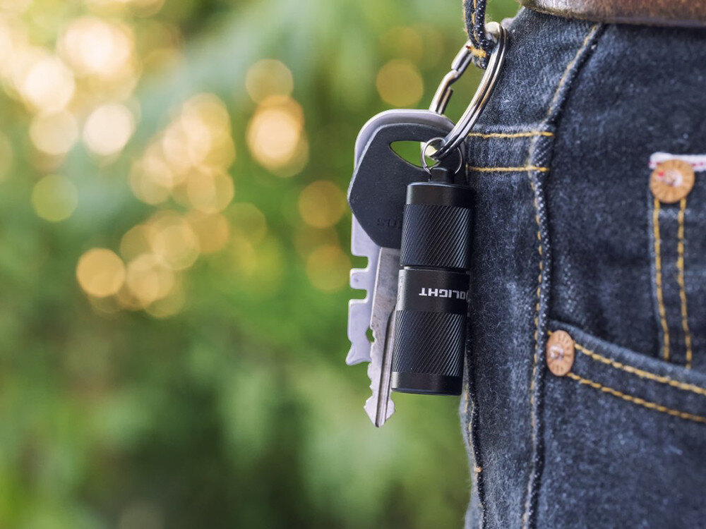 Latarka OLIGHT I1R II EOS Kit, brelok, metal, klucze, plecak, Czarna I1R 2 EOS zawieszona przy pasku spodni na breloku z kluczami, zielone rozmycie tła.