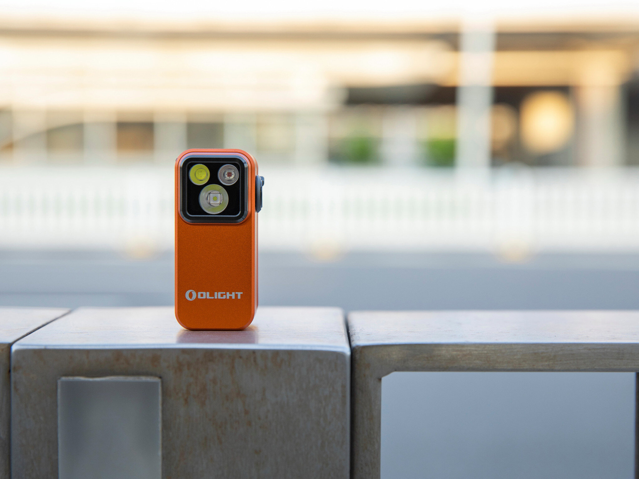 Latarka OLIGHT OL690834, trzy tryby, wszechstronna, lekka, do awaryjnych, Olight Oclip Pro pomarańczowa ustawiona na metalowej balustradzie, miejski rozmyty plener.