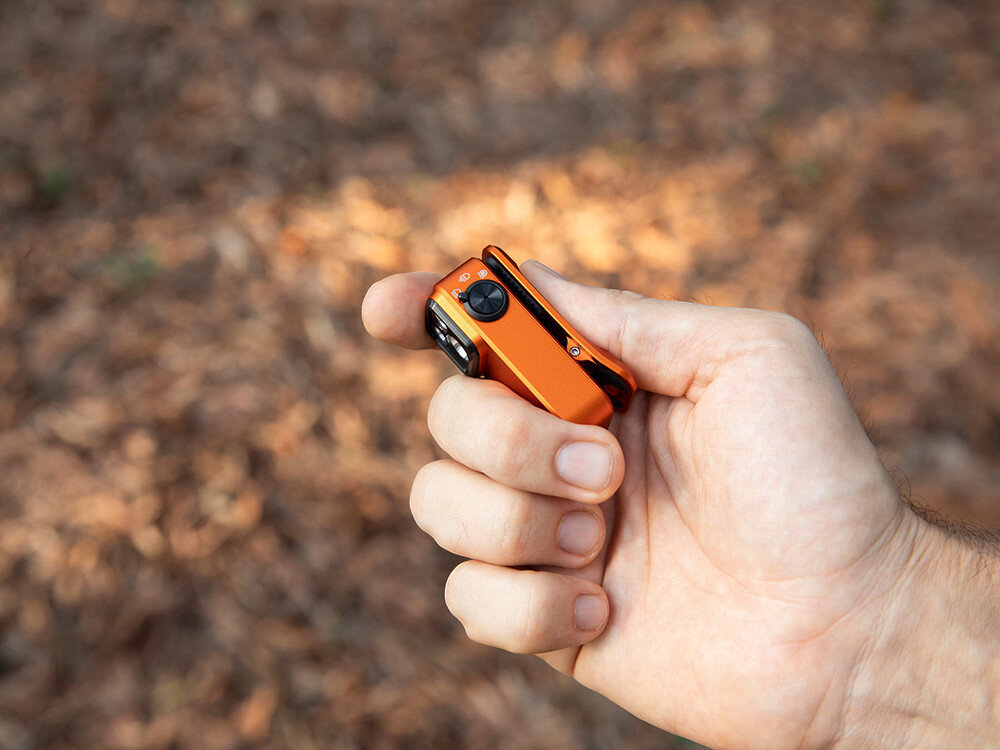 Latarka OLIGHT OL690834, aluminium, odporność, IPX6, outdoor, Oclip Pro trzymana na boku, prezentacja klipsa, aparat męska dłoń, las, jasne tło.