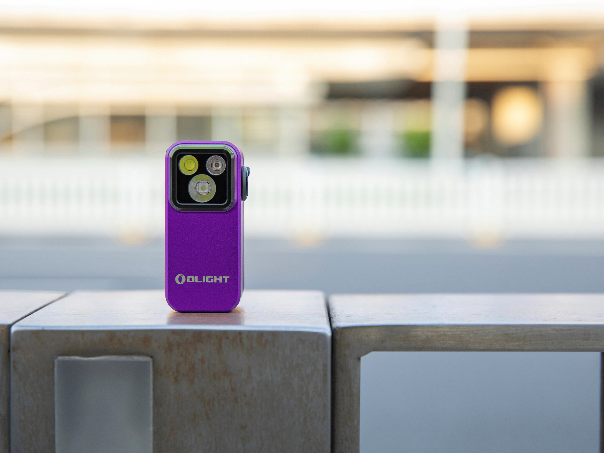 Latarka OLIGHT OL691572, trzy tryby, wszechstronna, lekka, do awaryjnych, Fioletowa latarka Oclip Pro na metalowej poręczy, rozmyte białe miasto w tle, design premium