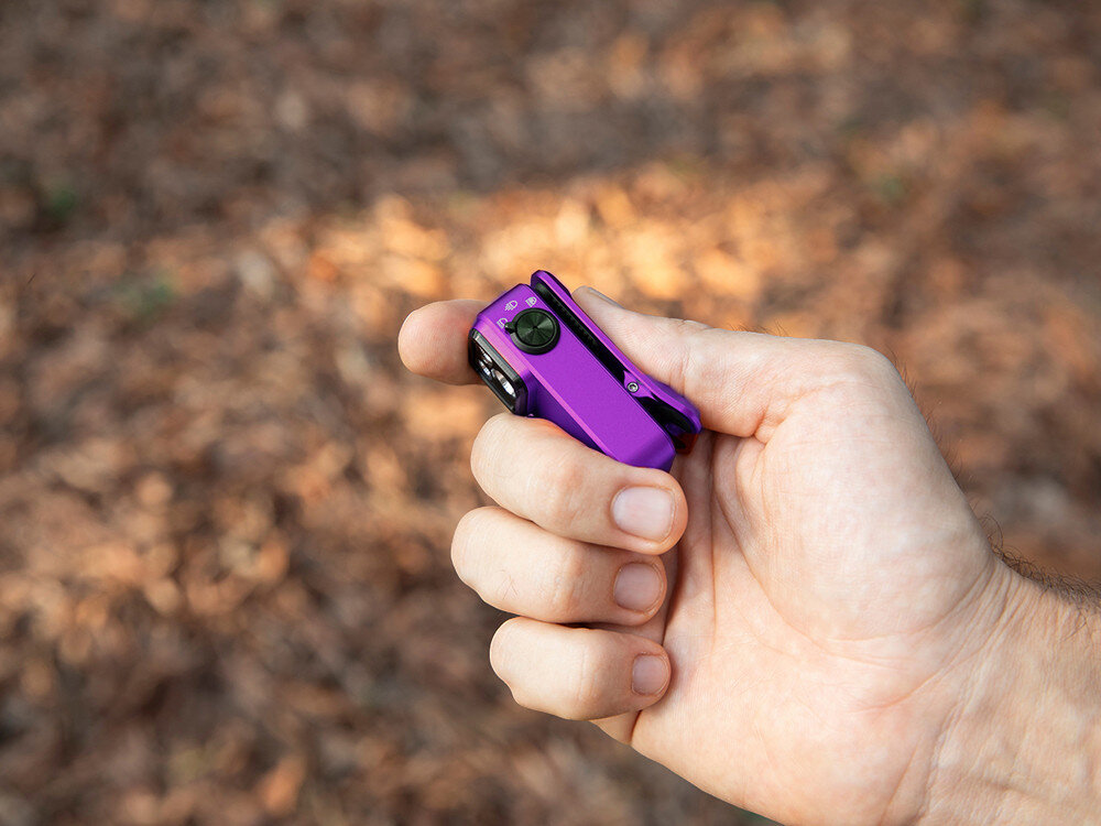 Latarka OLIGHT OL691572, aluminium, odporność, IPX6, outdoor, Fioletowa latarka Oclip Pro trzymana kciukiem od góry, detale przycisku i klipsa, liście w tle.