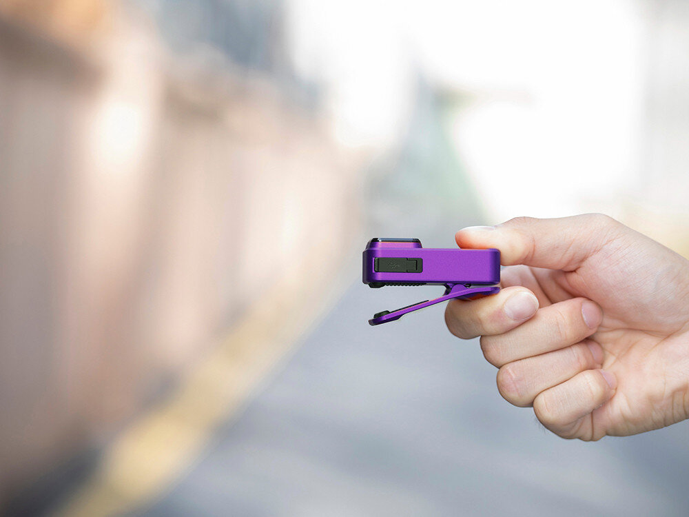 Latarka OLIGHT OL691572, magnes, klips, montaż, linka, Fioletowa Oclip Pro w dłoni bokiem, dobrze widoczny klips, jasny, miejski plener w tle.