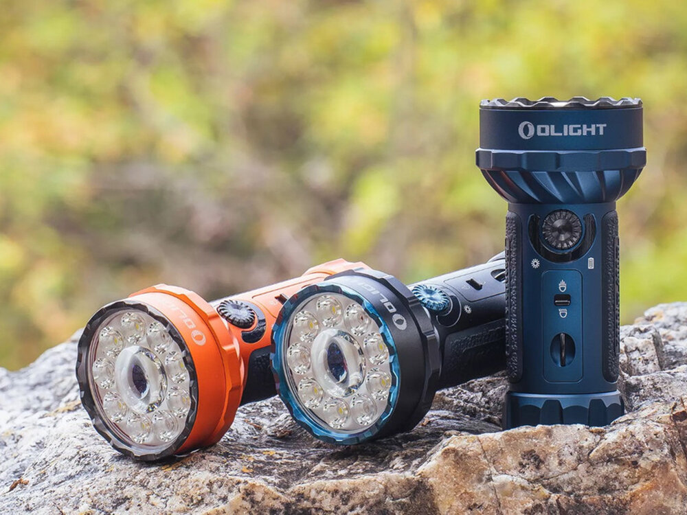 Latarka OLIGHT Marauder Mini QL000549 Niebieski, 7000 lumenów, ergonomia, floodlight, RGB, Czarno-niebieska latarka Olight Marauder Mini stoi pionowo na kamieniu, obok leżą wersje pomarańczowa i niebieska, ostre tło z zielenią.