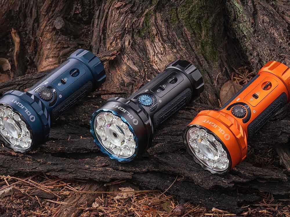 Latarka OLIGHT Marauder Mini QL000549 Niebieski, aluminium, IPX8, odporna, O-ring, Olight Marauder Mini w kolorach czarnym, niebieskim i pomarańczowym na tle pnia drzewa, leżą na ziemi w rzędzie.