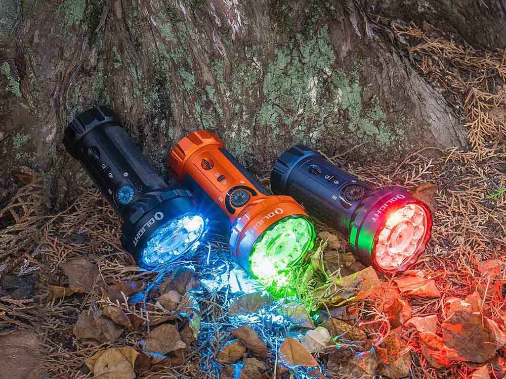 Latarka OLIGHT Marauder Mini QL000549 Niebieski, akumulator, ładowanie, wskaźnik, MCC3, Trzy latarki Olight Marauder Mini pod drzewem, każda świeci innym kolorem: niebieskim, zielonym, czerwonym.